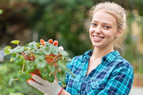 A young woman with blonde hair styled in a loose ponytail, dressed in a blue and green plaid shirt and white gardening gloves, is smiling as she holds a colourful pot with vibrant green foliage and small white flowers, standing outdoors in a garden setting. In the background, there are blurred trees and bushes, indicating a lush and well-maintained garden area. The scene is well-lit with natural daylight, suggesting a bright, clear day, and the overall environment appears tidy and inviting, reflecting typical features of a residential garden in Harrow, with a mixture of flowering plants, greenery, and natural textures suitable for gardening and outdoor landscaping activities by companies like Landscapers Harrow.