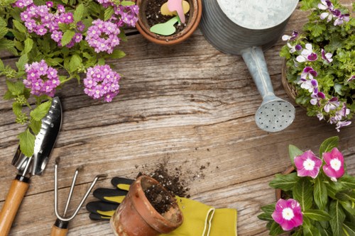 The image shows a wooden outdoor garden table with various gardening tools and plant containers arranged on it. There are flowering plants in pots, including pink and purple blooms, positioned around the table. A small terracotta pot with soil, a pair of black and yellow gardening gloves, and a yellow gardening trowel are visible, along with a metal hand rake. A grey watering can with a round head and a handle is placed at the top right. The scene suggests a setting for gardening or plant care, typical of a landscaped garden area in Harrow, with natural light indicating daytime weather. The table surface is weathered, showcasing the natural wood grain, while the background hints at a lush, outdoor environment suited for garden landscaping and flower bed design work by Landscapers Harrow.