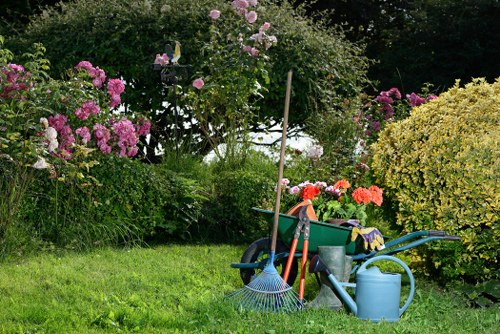 The image depicts a well-maintained garden area with a lush, green lawn occupying the foreground, bordered by a variety of flowering bushes and shrubs in shades of pink, purple, and yellow. In the background, taller trees provide a natural canopy, while a climbing rose is supported by a wooden stake. On the right side, there is a neatly trimmed hedge and a dense cluster of flowering plants, adding colour and texture to the scene. In the centre of the garden, several gardening tools and supplies are arranged on the grass, including a blue watering can, a green wheelbarrow filled with gardening equipment, a rake, a trowel, and a garden glove, indicating ongoing outdoor maintenance. The overall environment suggests a peaceful, private outdoor space ready for gardening care or relaxation, with natural daylight illuminating the scene, ideal for showcasing landscaping and gardening services, such as those offered by Landscapers Harrow.