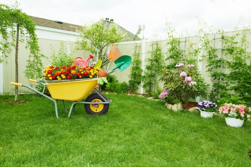 A well-maintained backyard garden with a lush, evenly cut green lawn set against a white fence. On the left side, a yellow wheelbarrow filled with colourful potted flowers, along with garden tools such as a watering can and small rake, are positioned on the grass. The background features a variety of shrubs and small trees, with some climbing plants along the fence. To the right, a flower bed contains blooming pink and purple flowers, with several white pots and containers arranged on the ground, showcasing a mix of flowering plants and greenery. The outdoor environment appears bright and clear, indicating a mild weather day, suitable for gardening activities. The layout demonstrates a neatly organized garden space ideal for outdoor care and planting, typical of services offered by landscapersharrow.co.uk in the Harrow area. This scene highlights key elements of garden maintenance, planting, and landscaping, emphasizing a vibrant and inviting outdoor landscape consistent with professional gardening services.