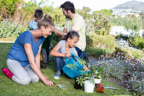 A family working together in a well-maintained garden with a lush green lawn, bordered by flower beds filled with blooming plants and shrubs. The garden features a neatly trimmed hedge along one side, providing structure and privacy, while a backdrop of trees and distant hills suggests a suburban setting near Harrow. The foreground shows a young girl planting new greenery, assisted by her mother who is kneeling on the grass, holding gardening tools and watering cans. Behind them, a father and son are engaged in gardening activities, with the father holding a small rake and the son using a trowel. The garden surface is predominantly grass with sections of paved pathways, and the scene is illuminated by natural daylight, indicating clear weather with vibrant colours and textures in the flora. The outdoor space appears to be cared for by professional landscaping services, highlighting the property's neat and inviting garden environment near Harrow, suitable for family outdoor activities and gardening projects.