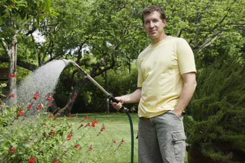 A man stands in a well-maintained garden with lush green grass and a variety of plants, watering flower beds with a garden hose. The garden features a neatly trimmed hedge in the background, alongside trees with fresh green foliage, suggesting a spring or early summer setting. The lawn appears dense and vibrant, bordered by a paved pathway or patio area, with a mix of flowering plants and shrubs close to the foreground. The scene is illuminated by natural daylight, highlighting the varied textures of foliage, soil, and paving. The man is dressed casually in a light yellow T-shirt and grey shorts, engaging in outdoor gardening activity, which aligns with landscaping and garden maintenance services offered by Landscapers Harrow in the Harrow area. This outdoor space demonstrates typical landscaping features such as lawn care, plant watering, and garden organization, indicating a professionally maintained residential garden designed for outdoor enjoyment and sustainable gardening practices. The overall environment is peaceful and inviting, showcasing a well-kept and vibrant garden scene.