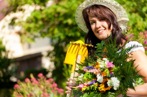 A woman in a garden during daytime, surrounded by lush greenery and flowering plants, holding a bright yellow gardening tool and a large bouquet of colorful flowers and foliage. The garden features a well-maintained lawn with dense, green grass in the foreground, bordered by flower beds with blooming flowers in shades of pink, purple, and white. In the background, there are trees and shrubs with varying heights, contributing to a peaceful outdoor environment. The woman is wearing a wide-brimmed hat, indicating sunny weather, and is smiling while engaging with the garden. The scene illustrates a healthy, landscaped outdoor space typical of residential gardens in Harrow, with natural textures of grass, soil, and plant foliage, subtly supporting local gardening and landscaping services by showcasing garden care and planting activities.