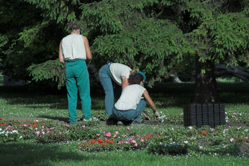 Three individuals are working together in a landscaped garden, surrounded by lush green trees and a well-maintained lawn. The person on the left, dressed in teal trousers and a sleeveless top, stands while observing. The other two, dressed in casual clothing, are kneeling and crouching near a flower bed featuring a variety of small, colorful flowers, including pink, red, and white blossoms, arranged in neat borders. The garden includes a mix of grass, flower beds, and patches of soil, with natural textures and vibrant colours, under soft daylight conditions. The scene depicts outdoor maintenance or planting activities typical of professional gardening services in Harrow, with a focus on sustainable landscaping practices that support local environmental efforts.