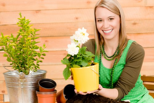 The image shows a woman with light brown hair smiling outdoors, sitting in a garden area with a wooden fence in the background. She is holding a small yellow pot containing a flowering plant with white blossoms and green leaves. In front of her on a wooden surface, there are additional gardening items, including a metallic pot with a leafy shrub, a black plastic pot, and empty terracotta pots. The garden scene features a well-maintained lawn with dense, green grass, and nearby plantings or shrubs are visible. The environment appears to be a neatly landscaped backyard or front garden, likely in Harrow, with natural light indicating a bright day. This setting reflects typical outdoor gardening activities, such as planting and potting, and aligns with services provided by Landscapers Harrow specializing in outdoor garden renovation and maintenance.