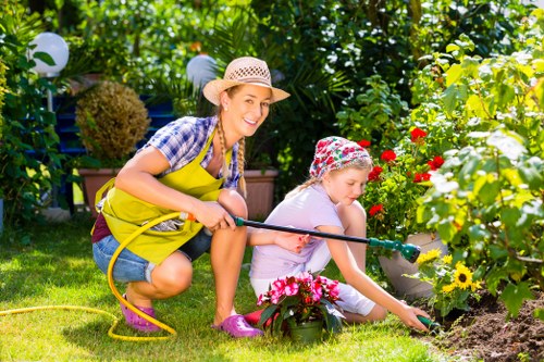 In a colorful garden, an adult woman and a young girl are engaged in outdoor gardening activities. The woman, dressed in a checked shirt, yellow apron, and wide-brimmed straw hat, is smiling while holding a yellow garden hose. The girl, wearing a pink and white floral hat and casual clothes, is kneeling on the grass and watering a flower bed with a small watering can. The garden features a well-maintained lawn with lush, green grass, a flower bed bordered by stones, and a variety of plants, including vibrant red, pink, and yellow flowers. In the background, there are leafy shrubs, trees, and garden decorations such as white spherical garden lights. The scene depicts a bright, sunny day with natural light highlighting the vibrant colours and healthy plants, illustrating a well-tended outdoor space suitable for family gardening and landscaping service improvements in the Harrow area, as referenced on the landscapersharrow.co.uk website for gardening and outdoor maintenance.