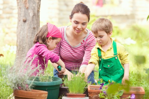 In a bright outdoor garden setting, a woman with dark hair tied back is engaging with two children, a girl wearing a pink headband and a yellow jacket, and a boy in a green apron. They are gathered around a variety of potted plants and garden tools, indicating active gardening or planting activity. The garden features a lush, green lawn with a large tree trunk visible on the left side, and blooming plants add colour and natural textures to the space. In the background, a garden fence and soft sunlight filtering through trees suggest a pleasant, well-maintained outdoor environment typical of residential properties in Harrow. The scene reflects a focus on nurturing garden growth and sustainability, consistent with the gardening services provided by Landscapers Harrow in the local area.